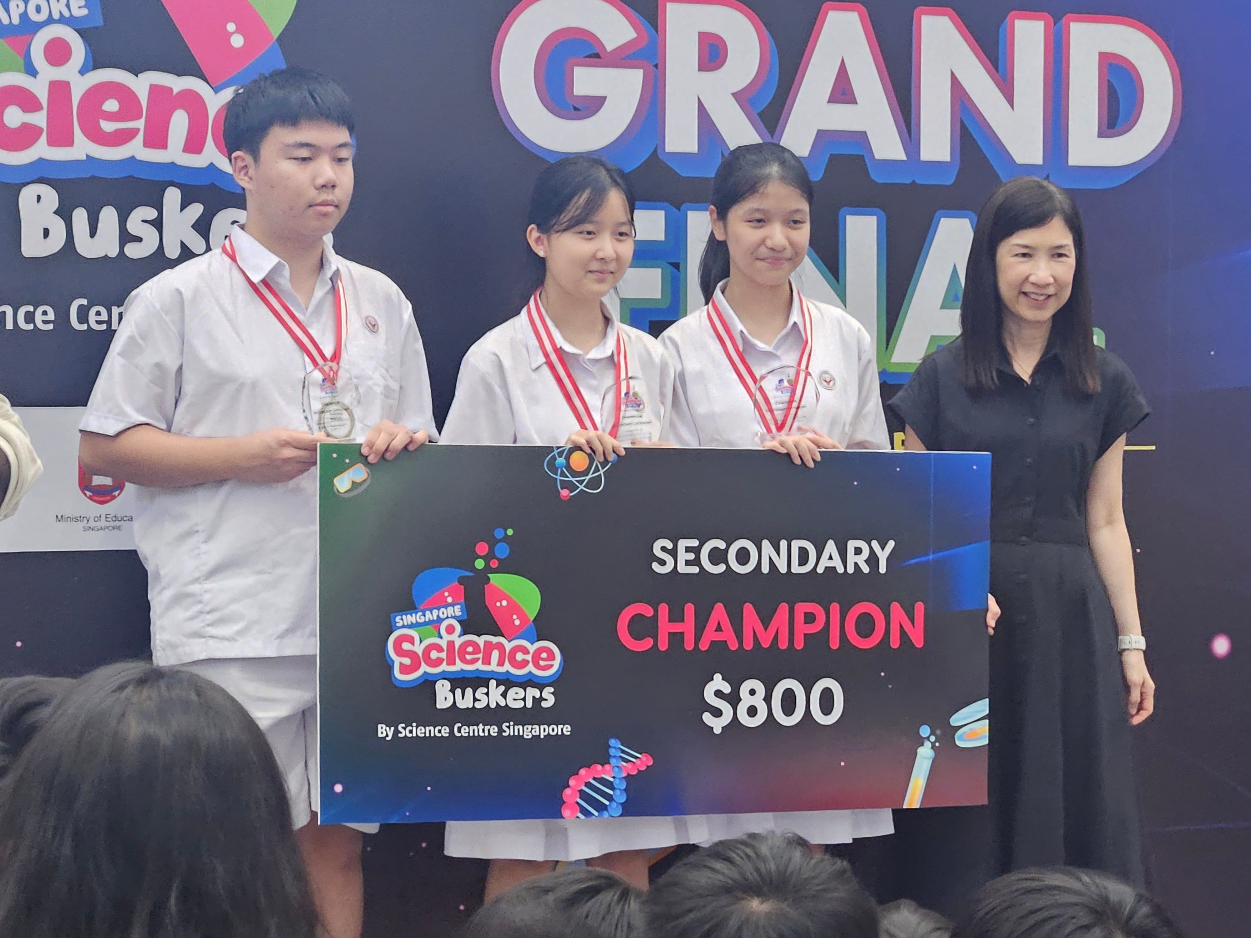 Three students with medals hold a Singapore Science Buskers Secondary Champion $800 prize board at a Grand Final event.