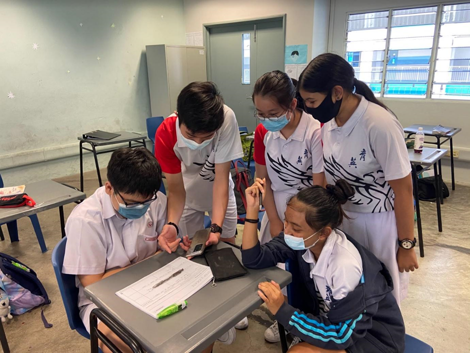 Five masked students in school uniforms gather around a desk, looking at papers and a phone in a classroom.