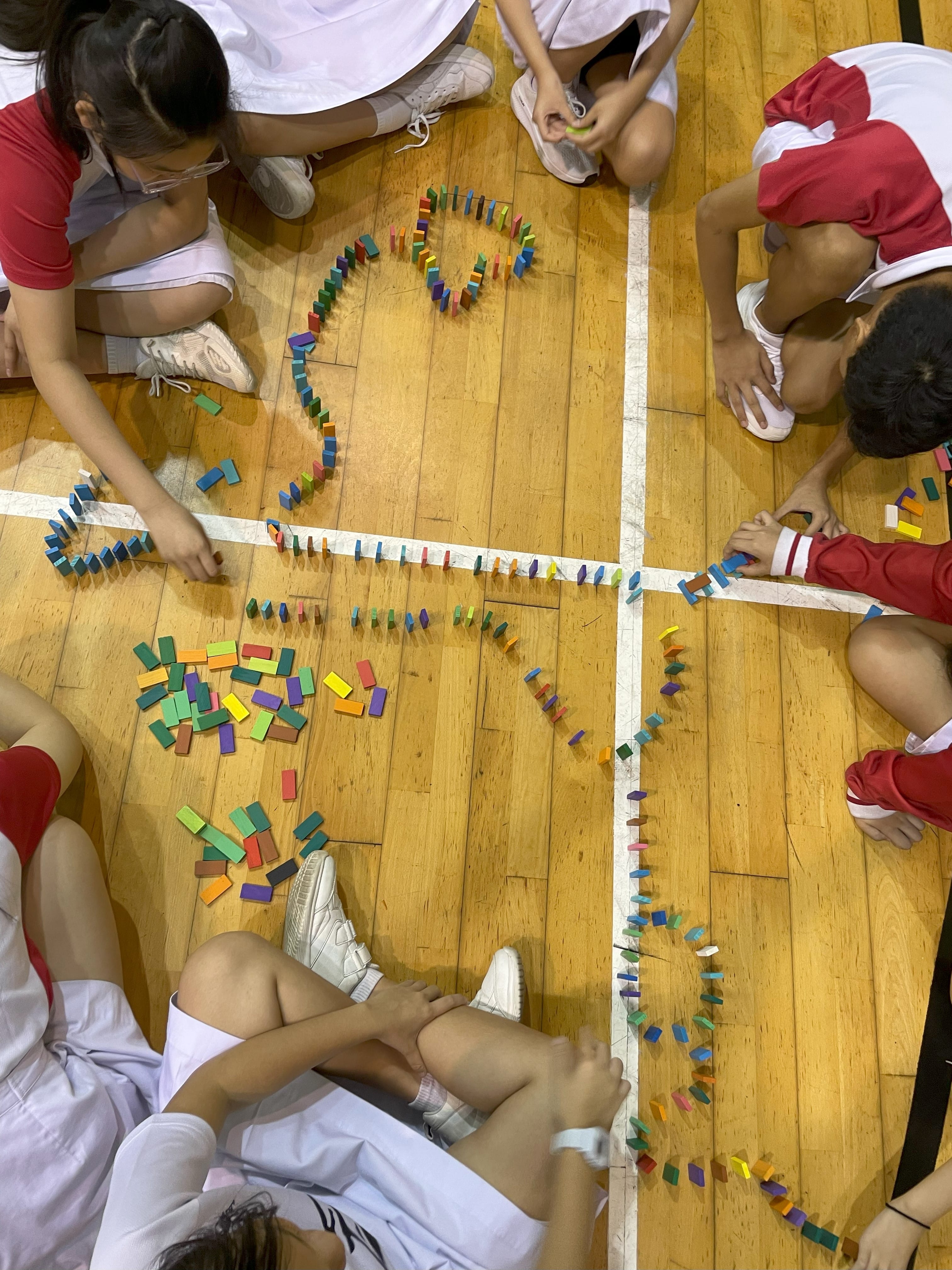 Children sitting on a wooden gym floor arranging colorful dominoes in long winding lines and a heart shape.