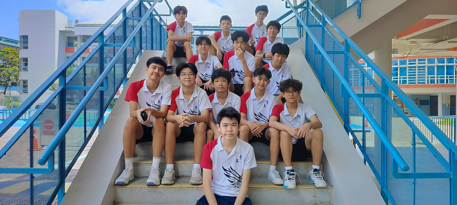 A group of teenage boys in matching white and red school uniforms pose on outdoor stairs with blue railings.