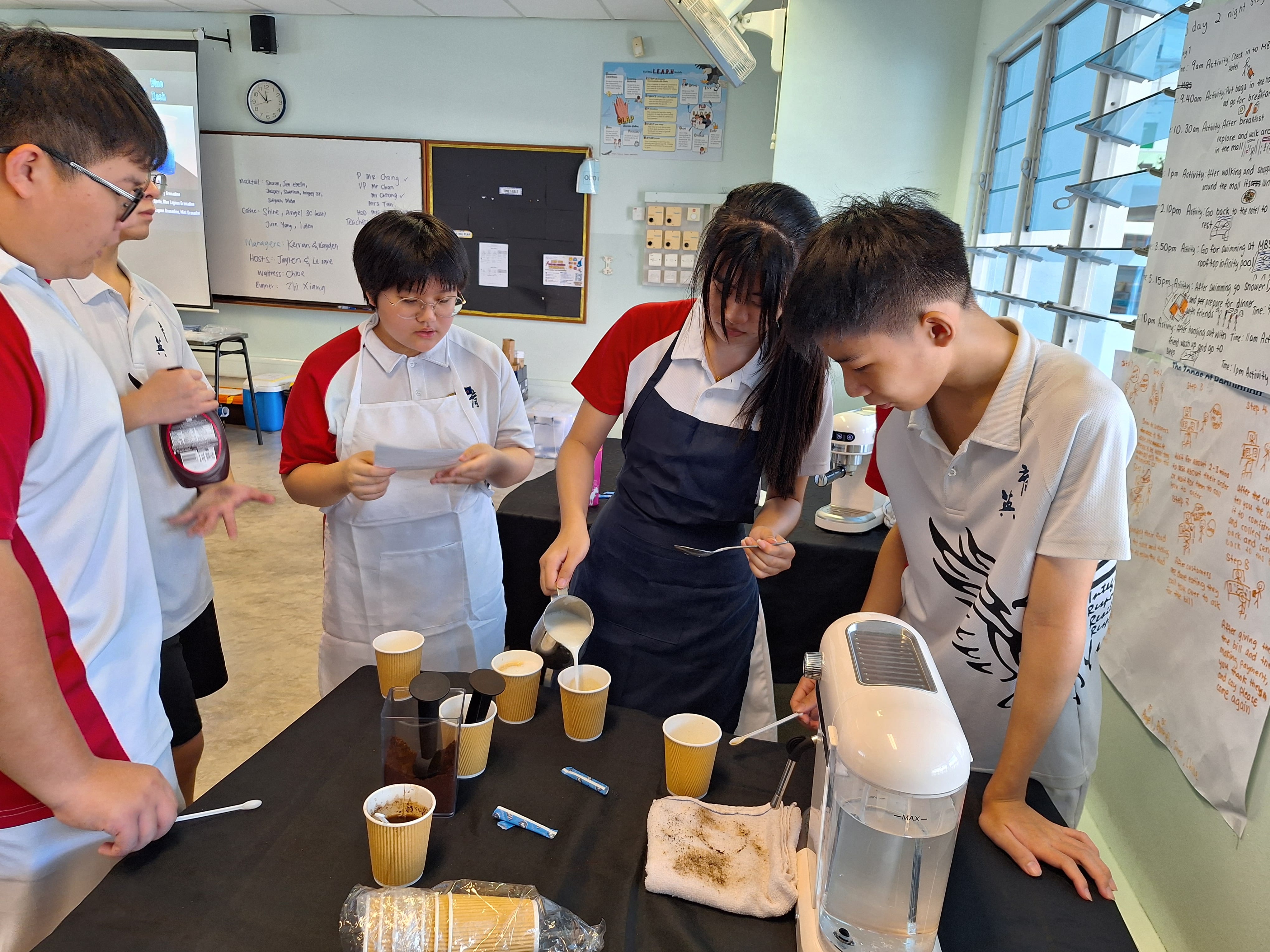Four students in school uniforms and aprons practice making coffee drinks at a table with cups and a machine in a classroom.