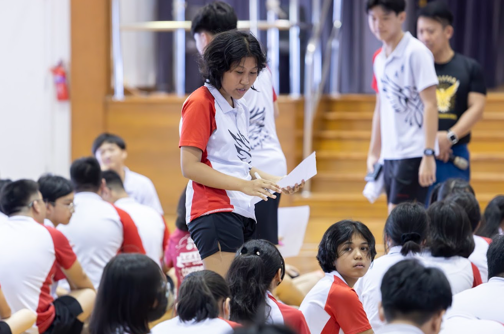 A girl in a red and white uniform holds papers while standing among students seated on a gym floor.