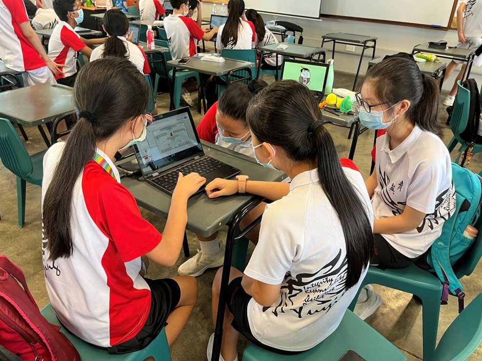 Three masked schoolgirls in uniforms gather around a laptop at a desk in a busy classroom.