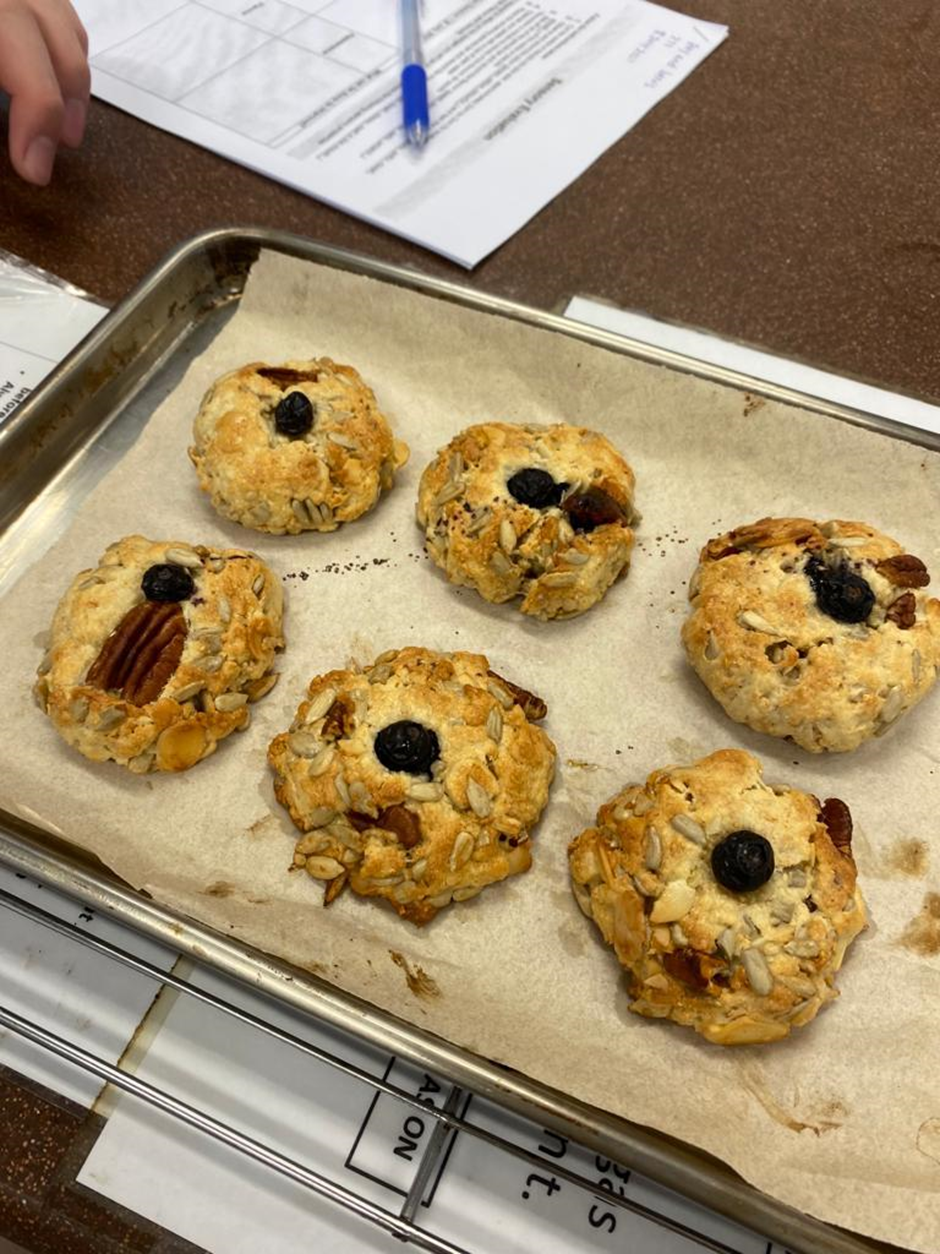 Six baked scones topped with sunflower seeds, nuts, and blueberries on a parchment-lined metal baking tray.