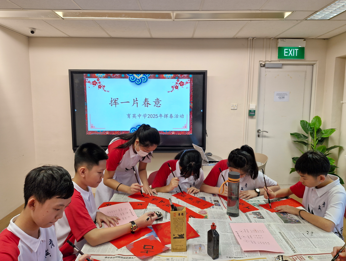 Students in school uniforms practice Chinese calligraphy on red paper at a table, with a festive Chinese slide shown behind them.