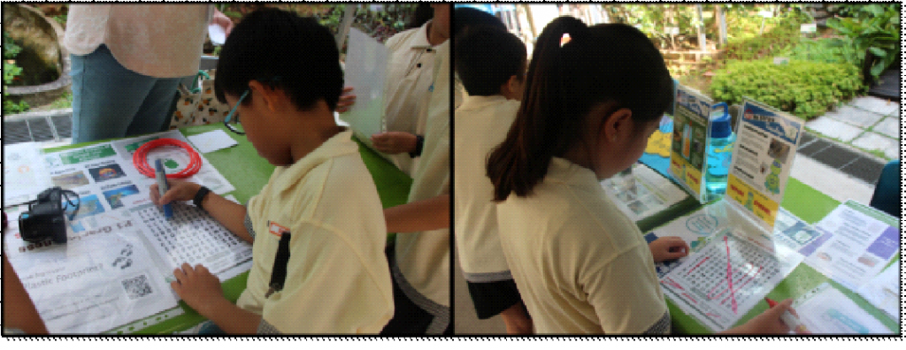 Two children in yellow shirts writing on large activity sheets at a table outdoors near green trees.