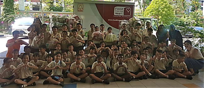 A large group of schoolchildren in uniforms pose with heart-hand gestures in front of a Food From the Heart truck.
