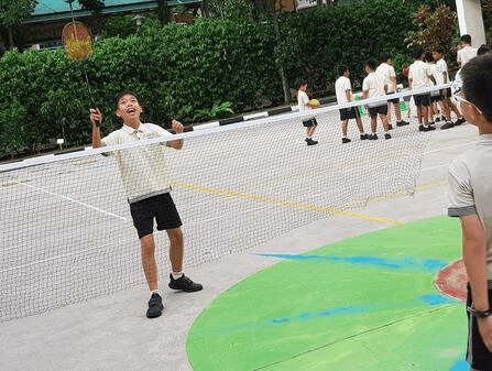 A smiling boy holds a badminton racket near a net on an outdoor court with other students in the background.