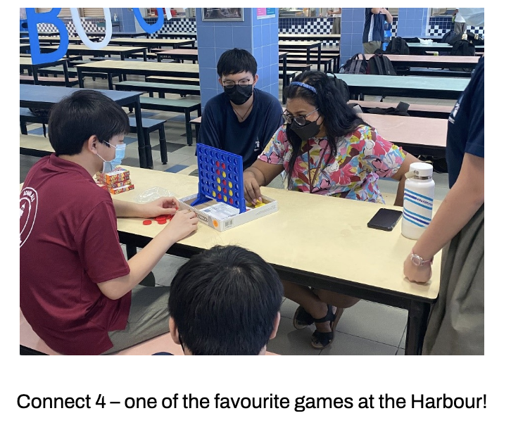 Our students sit around a cafeteria table playing Connect 4 board game with a teacher. 