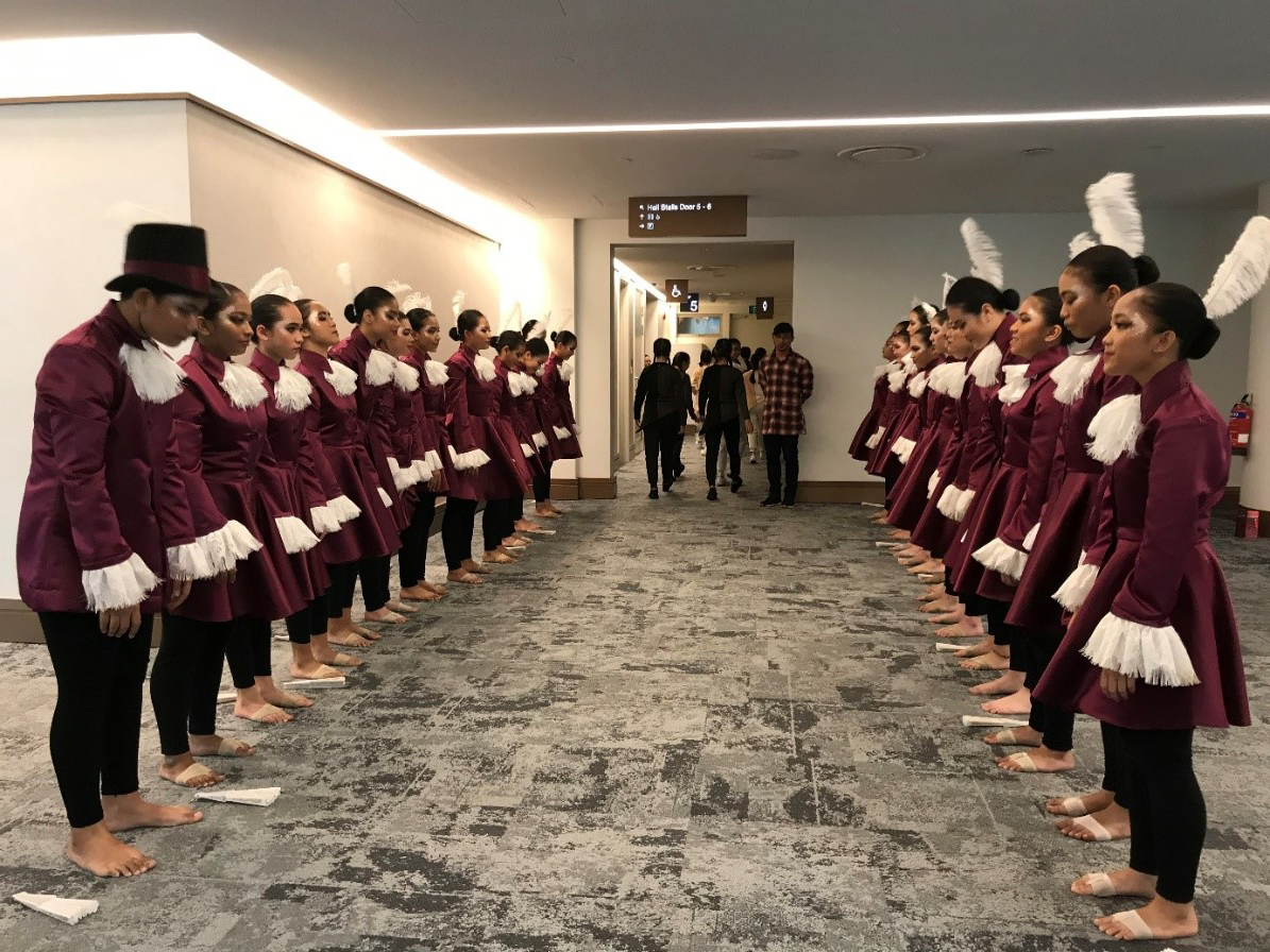 Two rows of performers in matching maroon jackets with white feather trim stand facing each other in a hallway.