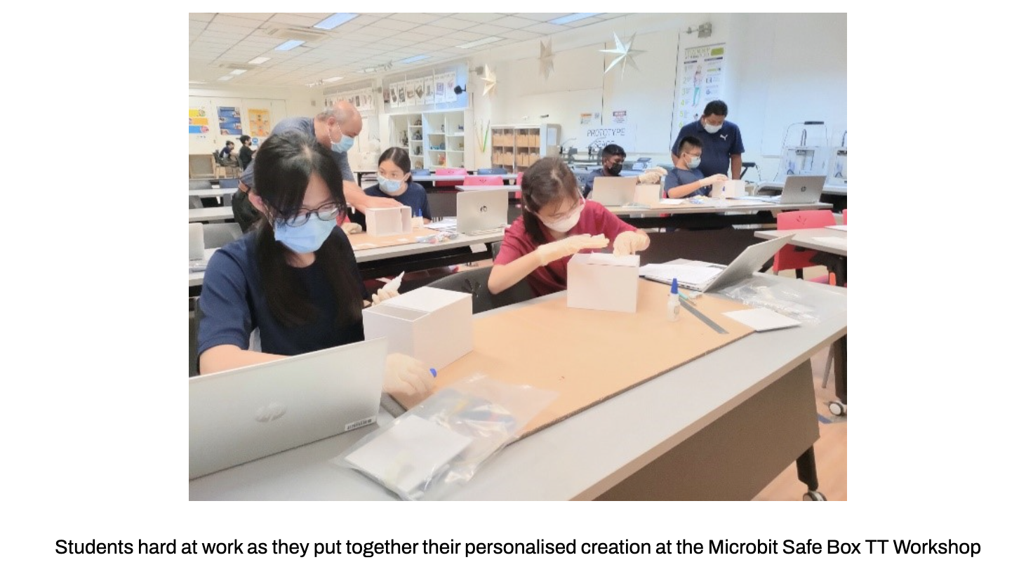 Students working on small white boxes with laptops open at desks in a bright classroom.