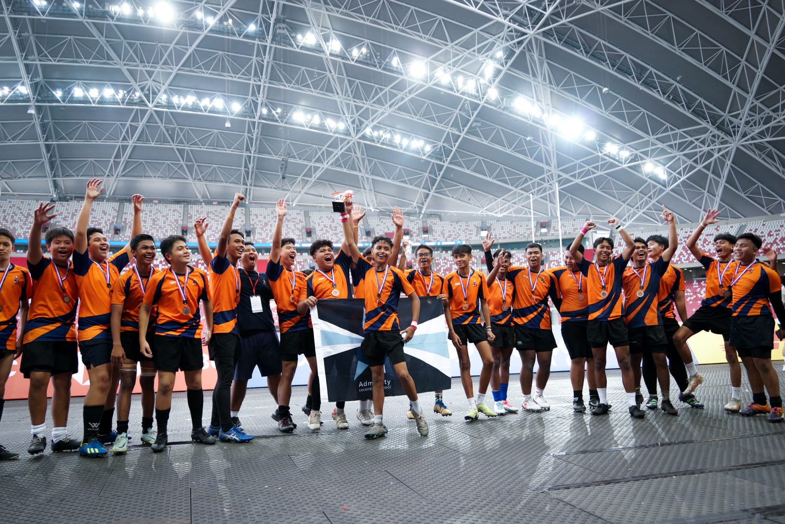 A youth rugby team in orange jerseys celebrates with medals and a trophy inside a large indoor stadium.