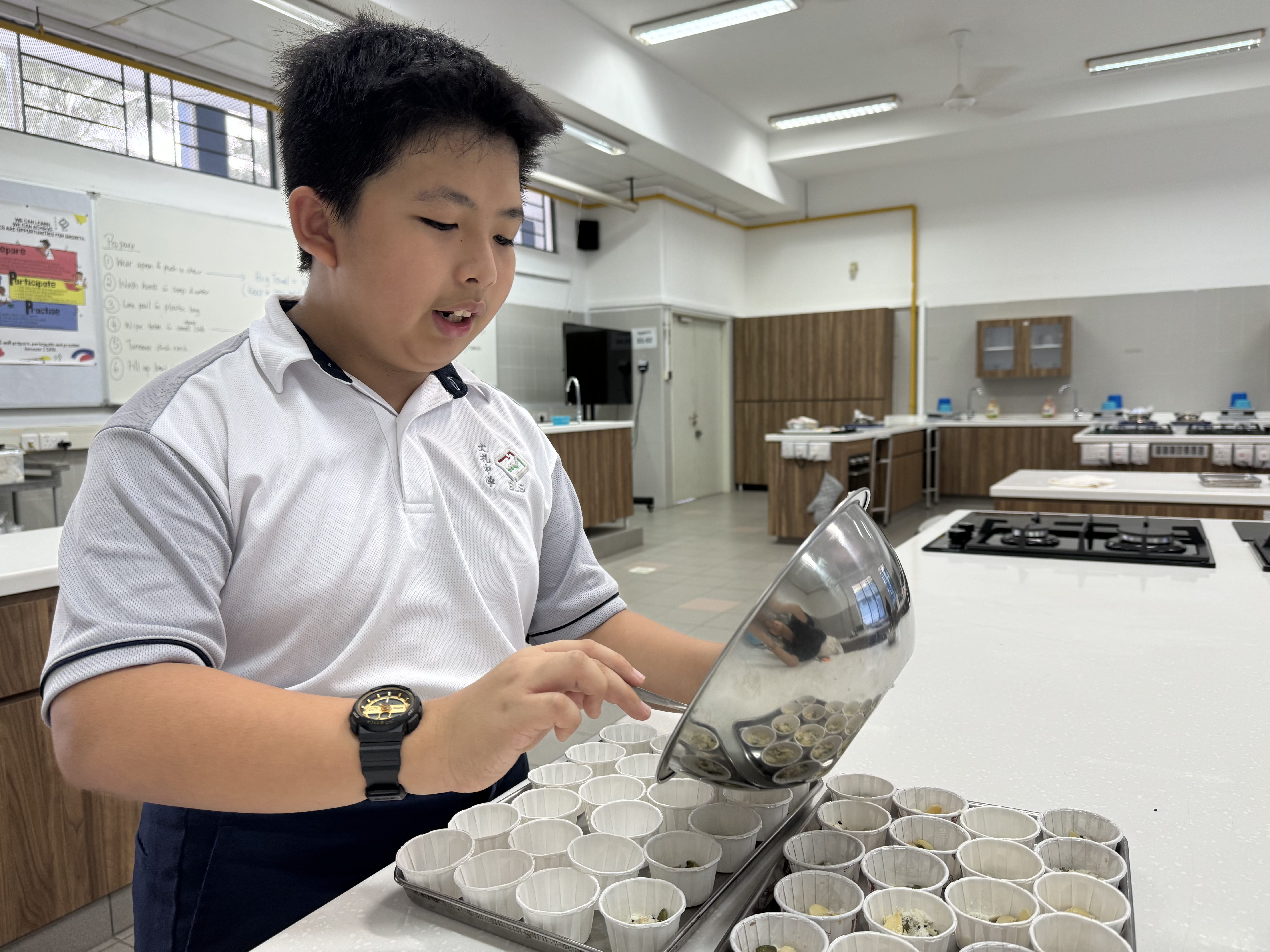 Student in a kitchen classroom pours batter from a metal bowl into many small paper cups on trays.
