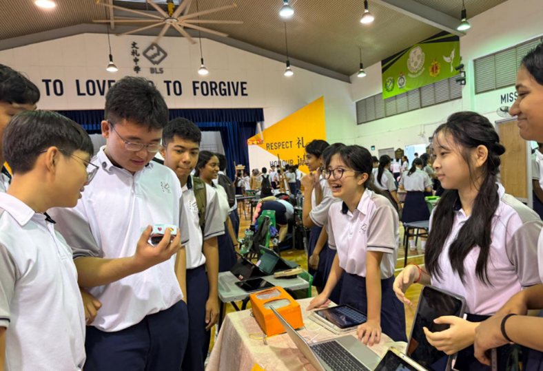Students in school uniforms gather around a display table with a laptop and devices in a school hall.