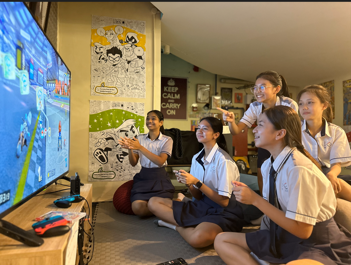 Five girls in school uniforms sit together, smiling and playing Mario Kart on a large TV.