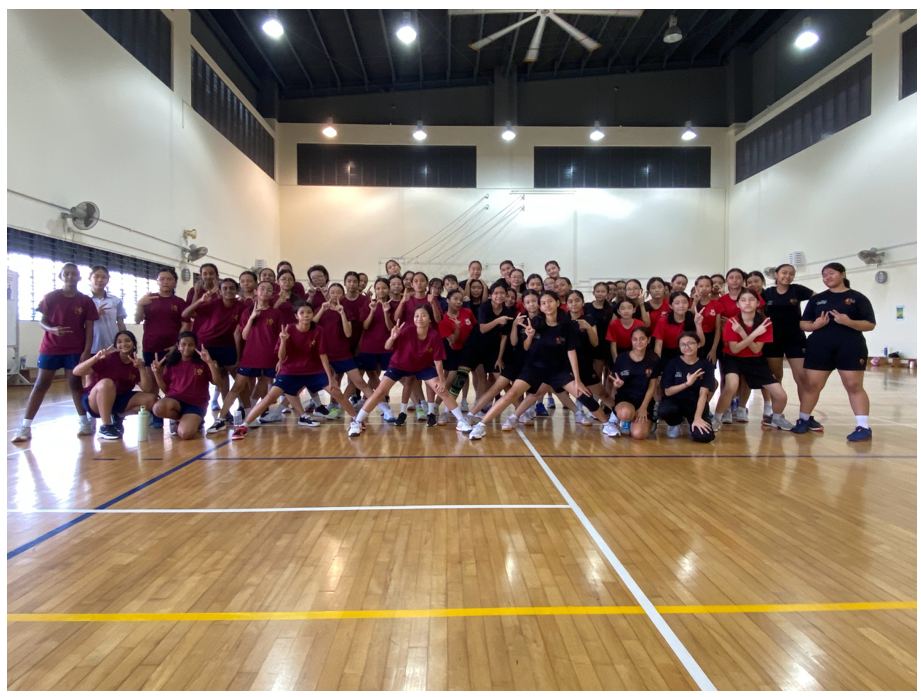 Large group of young people in red and black sportswear posing together inside an indoor wooden-floored gymnasium.
