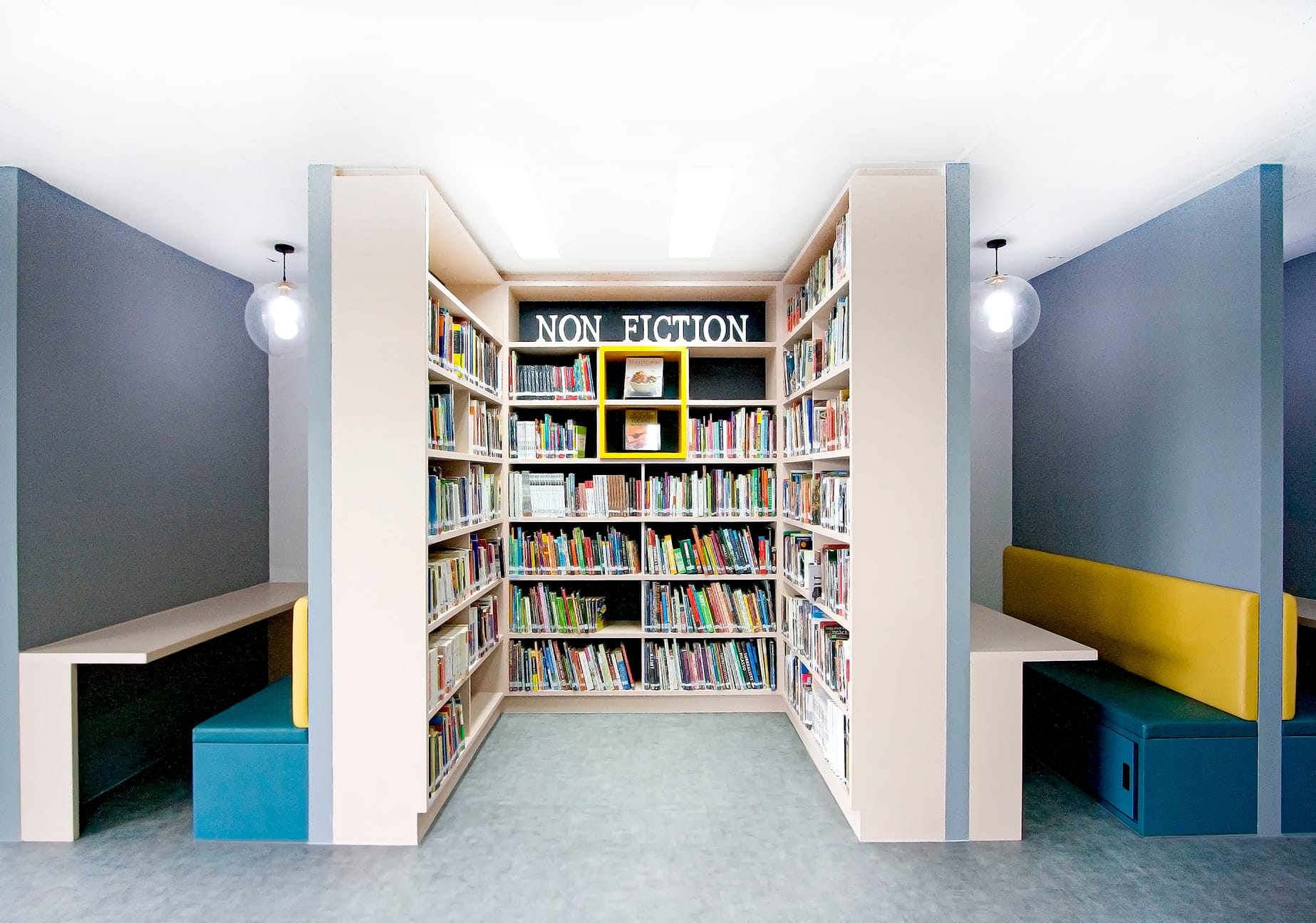 Library non fiction section with tall shelves full of books, yellow display cubes, and seating nooks on each side.