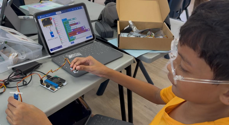 A boy wearing safety goggles works with a micro:bit circuit board connected to wires beside a tablet showing block coding.