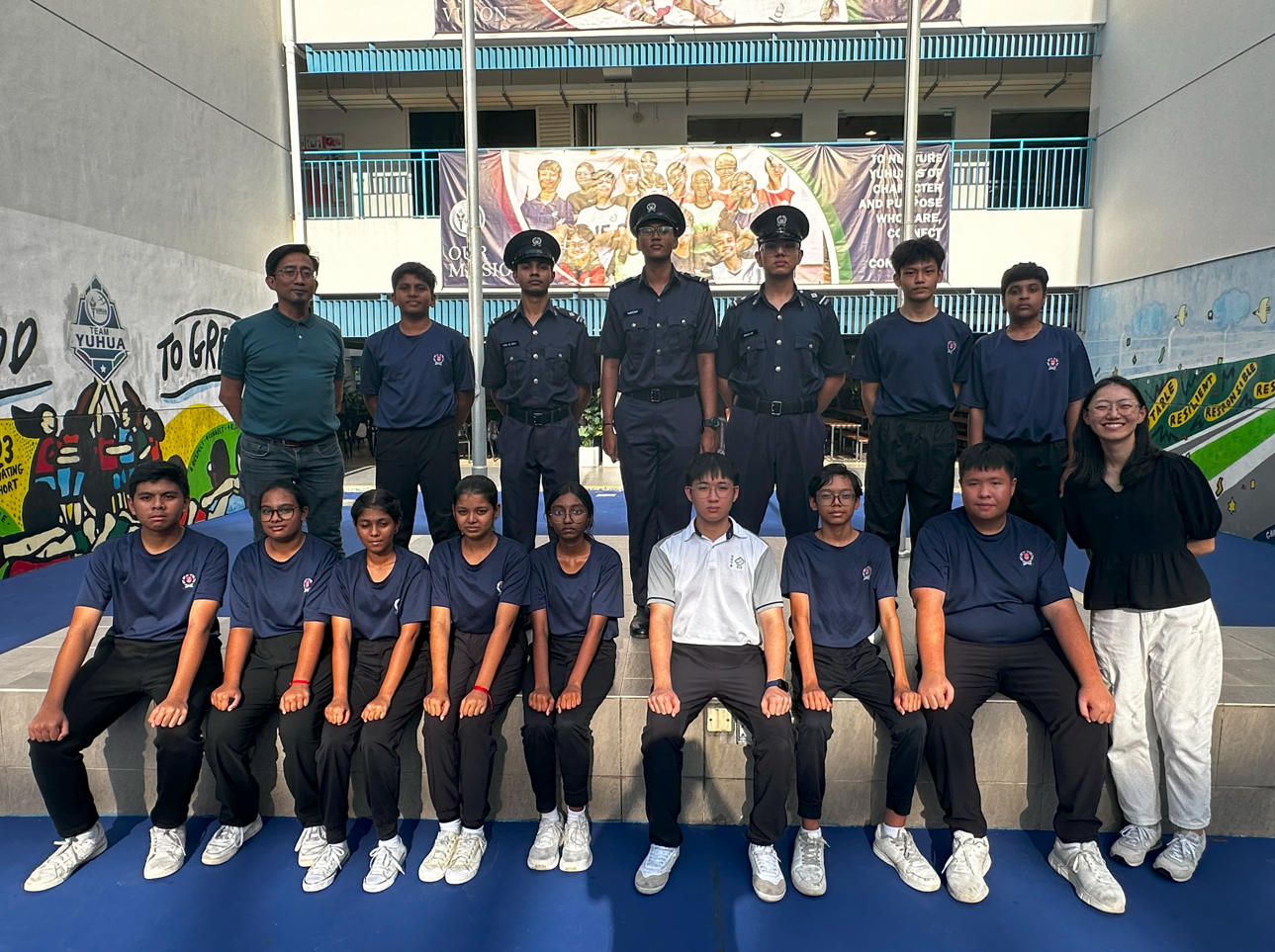 A group of students in navy uniforms and some in dark cadet uniforms pose in two rows at a school courtyard.