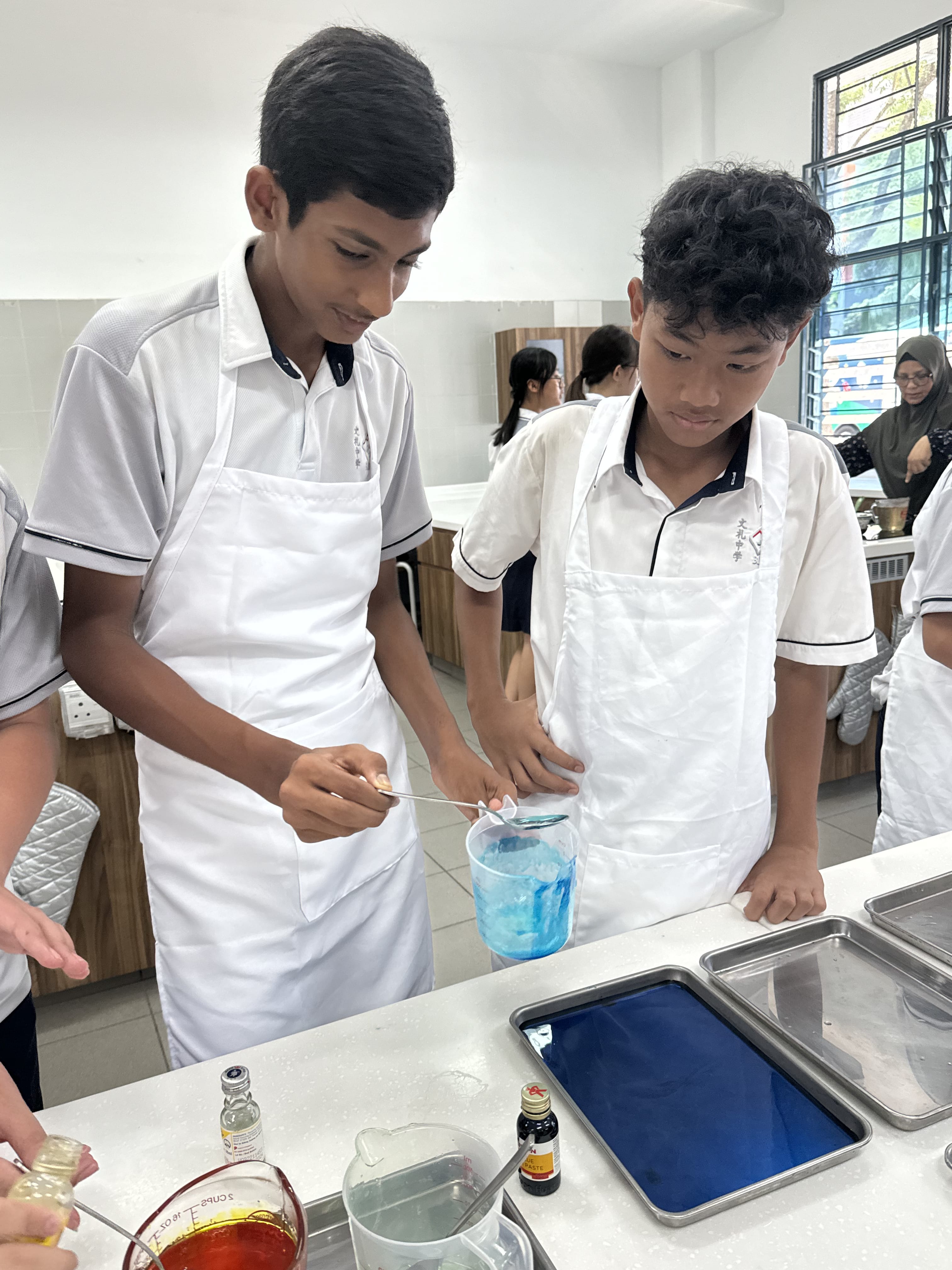 Two boys in white aprons mix blue liquid in a cup during a school cooking class.