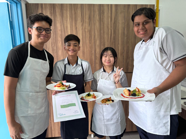 Four students in aprons hold plated dishes and a certificate, smiling in a kitchen setting.