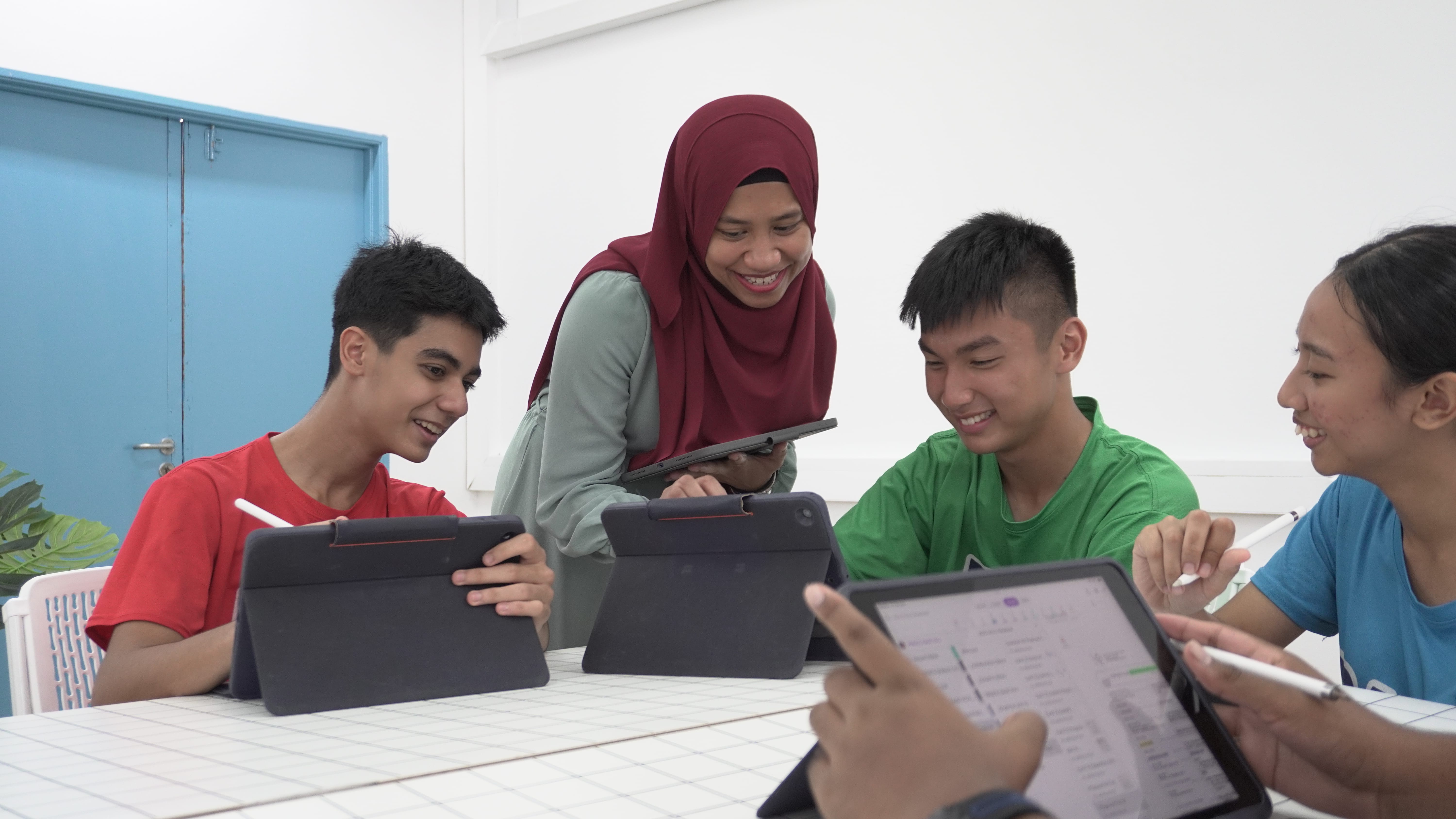 Teacher smiles while students use tablets at a classroom table.