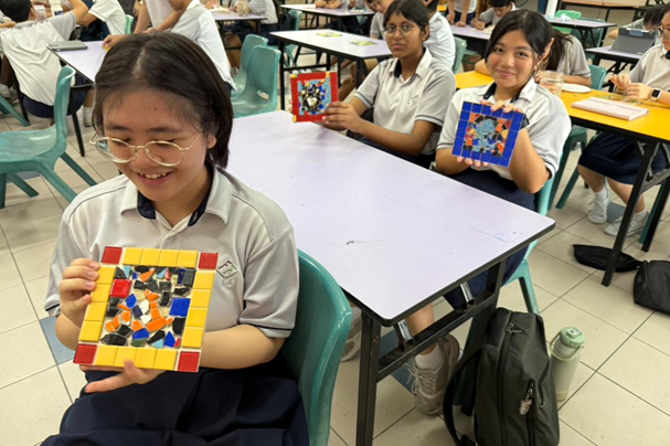 Three smiling schoolgirls in uniforms hold up colorful mosaic tile artwork in a classroom.
