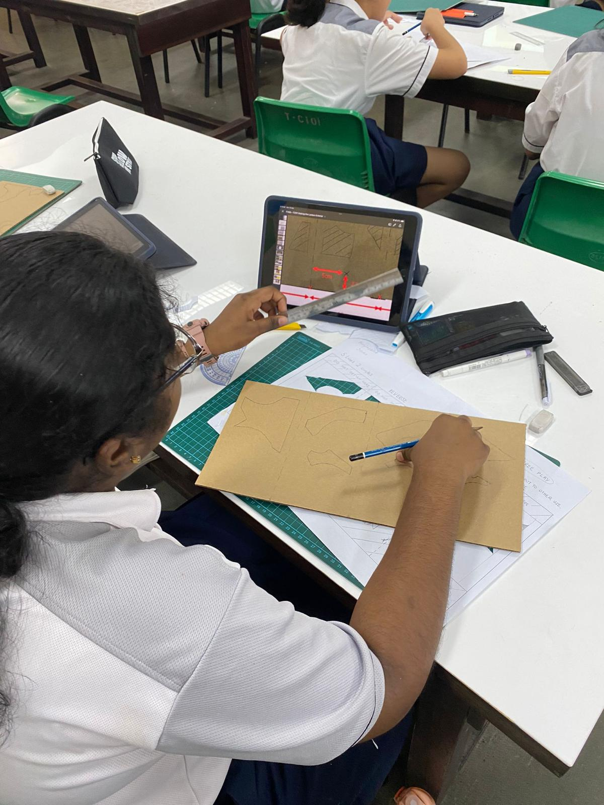A student in uniform draws geometric shapes on cardboard at a desk, referencing a tablet displaying a design tutorial.