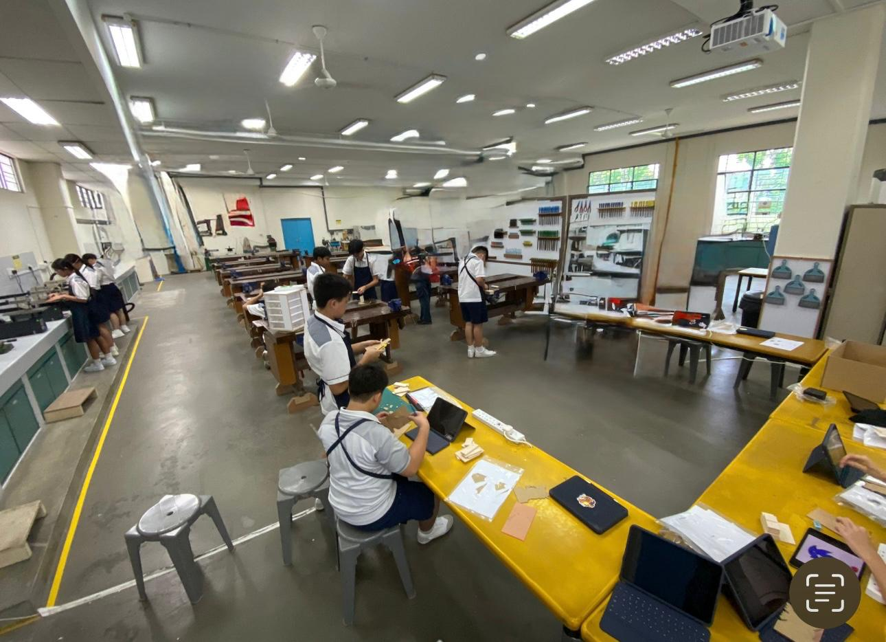 Students in school uniforms work at wooden benches in a large workshop classroom with tools on the walls.