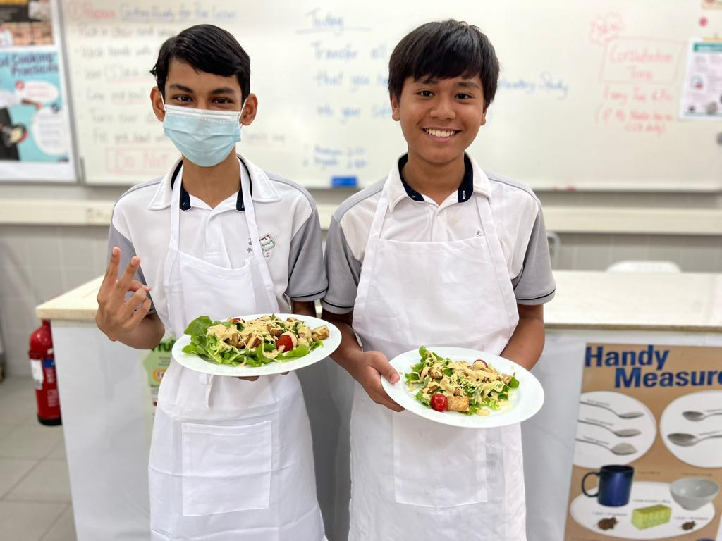 Two young students in white aprons hold plates of salad in a classroom, one wearing a face mask.