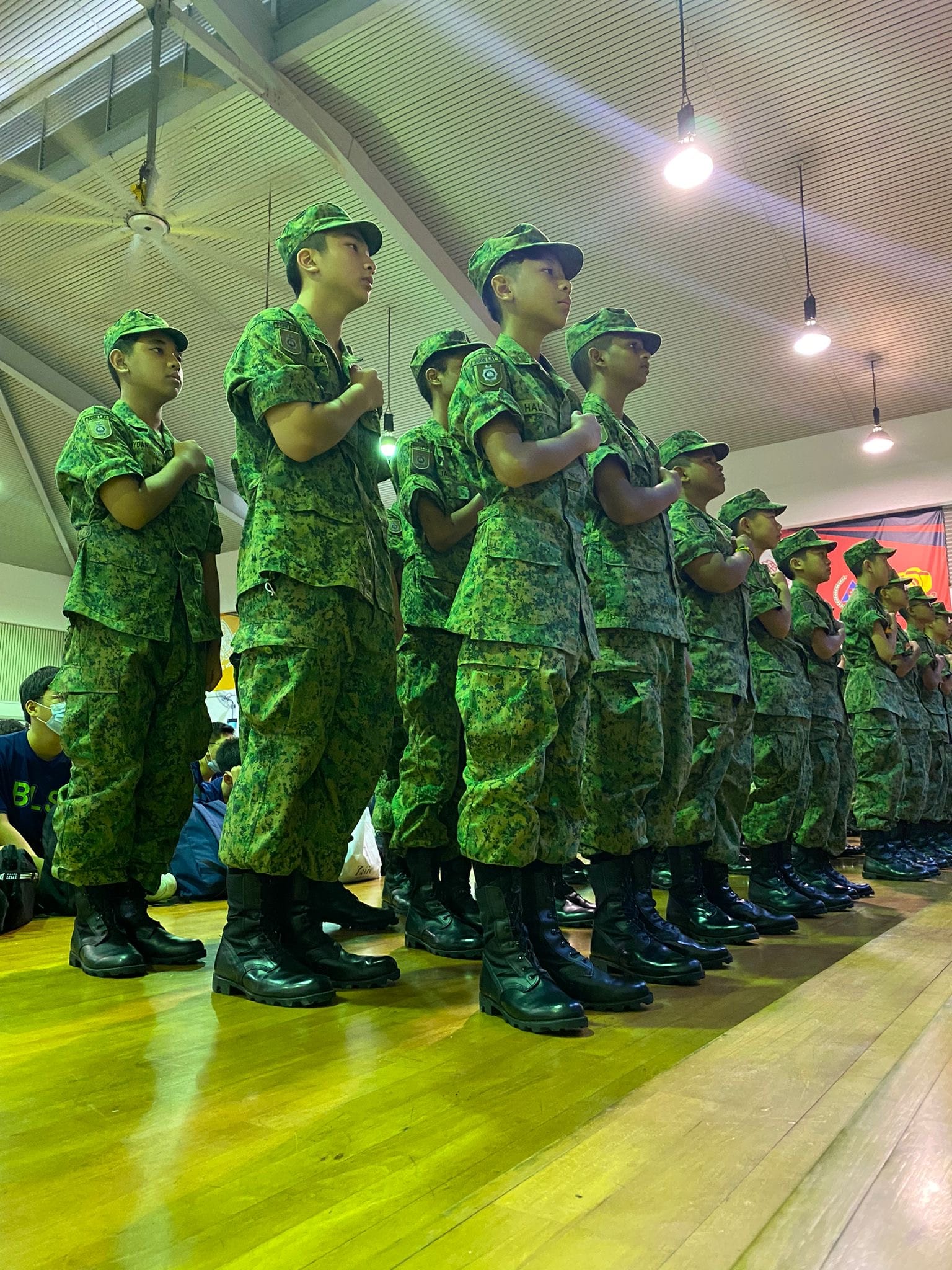 Rows of young cadets in green camouflage uniforms and caps standing at attention on a wooden floor indoors.