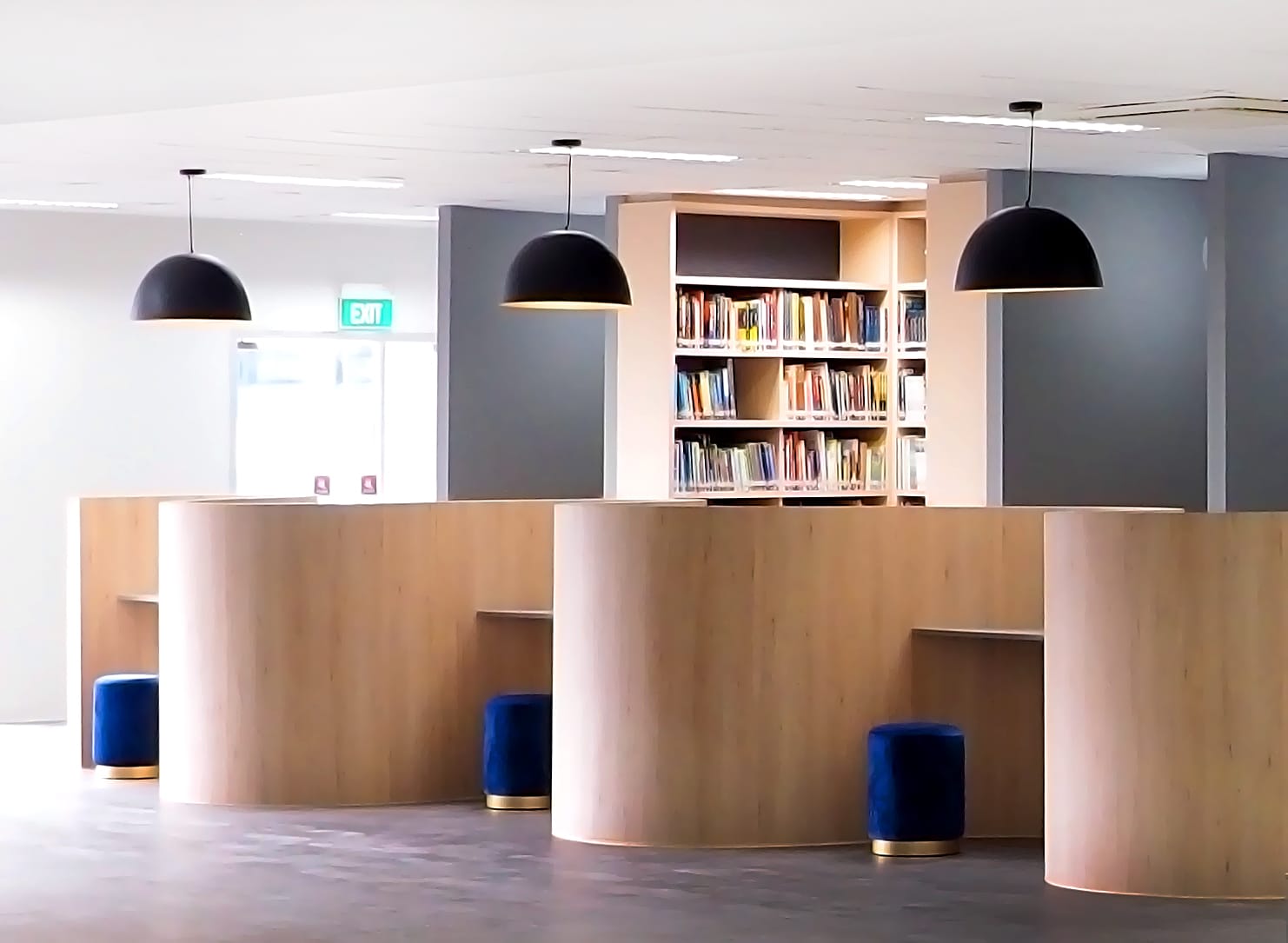 Modern library interior with curved wood desks, blue stools, black pendant lights, and a bookshelf filled with books.