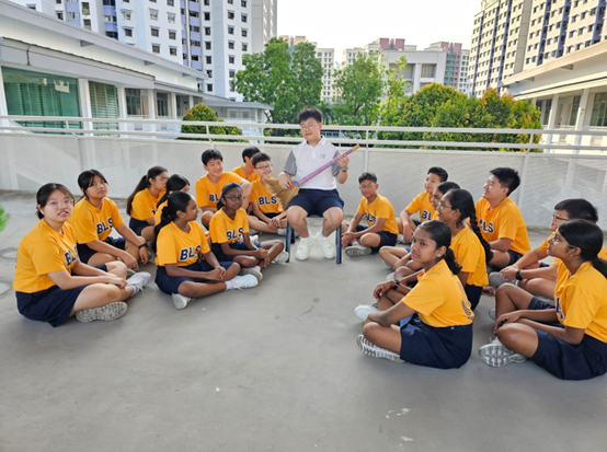 A teacher holds a pink object while students in yellow BLS shirts sit in a circle on an outdoor rooftop area.