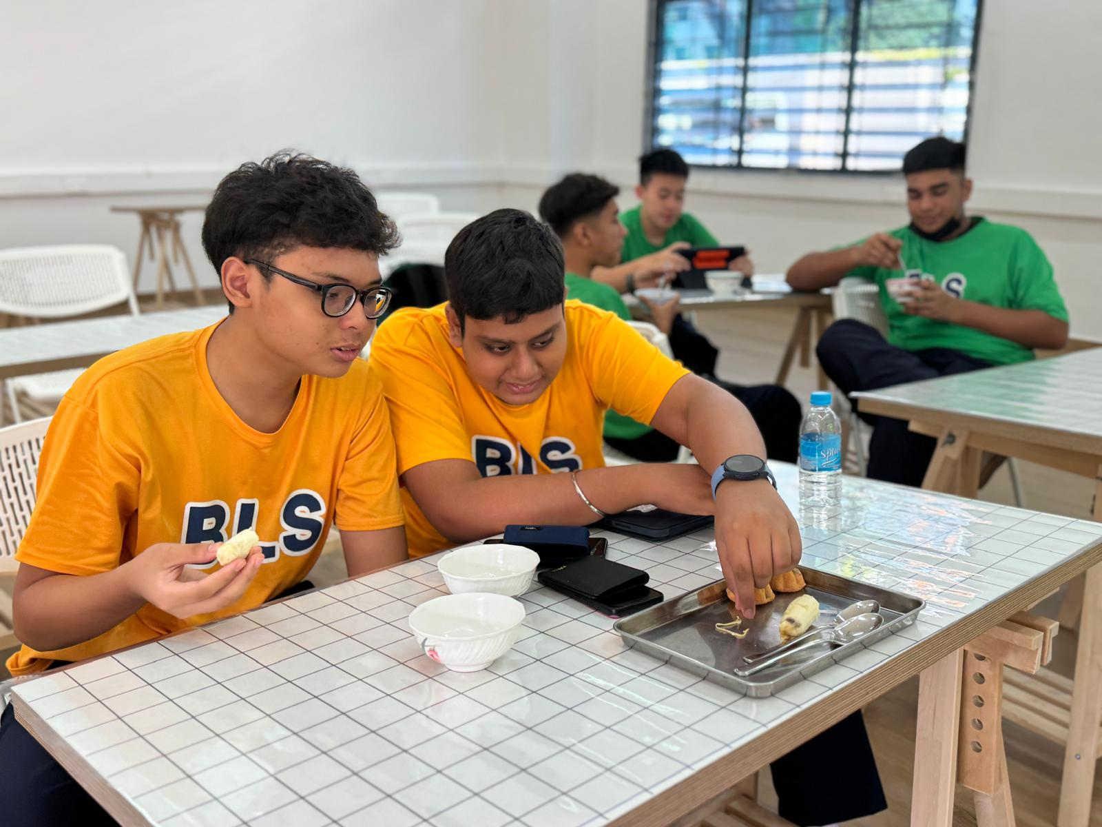 Two boys in yellow BLS shirts eat snacks at a tiled table; other teens in green shirts sit in background.