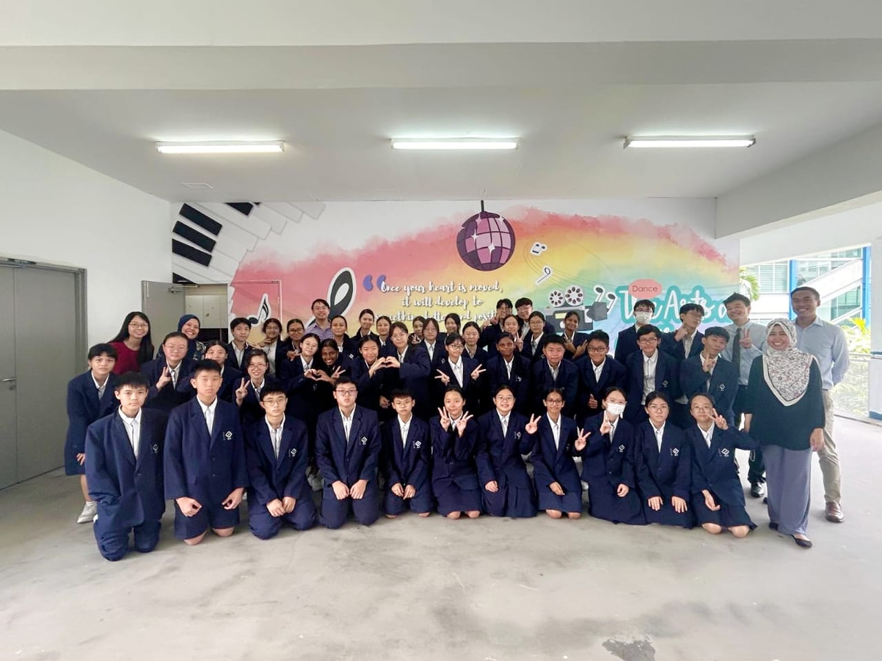 A large group of students in navy blazers pose in front of a colorful music-themed mural with teachers.