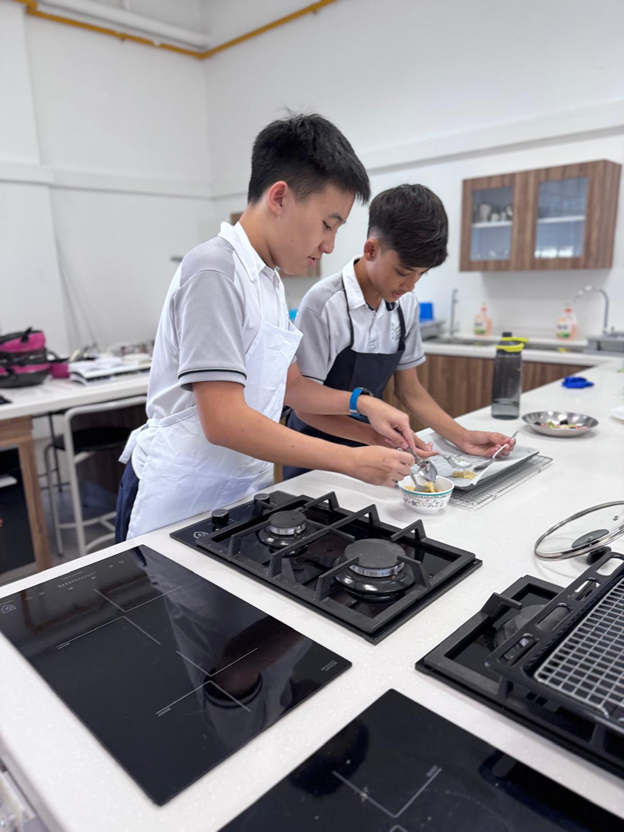 Two young male students in aprons prepare food at a kitchen counter with gas and induction cooktops nearby.