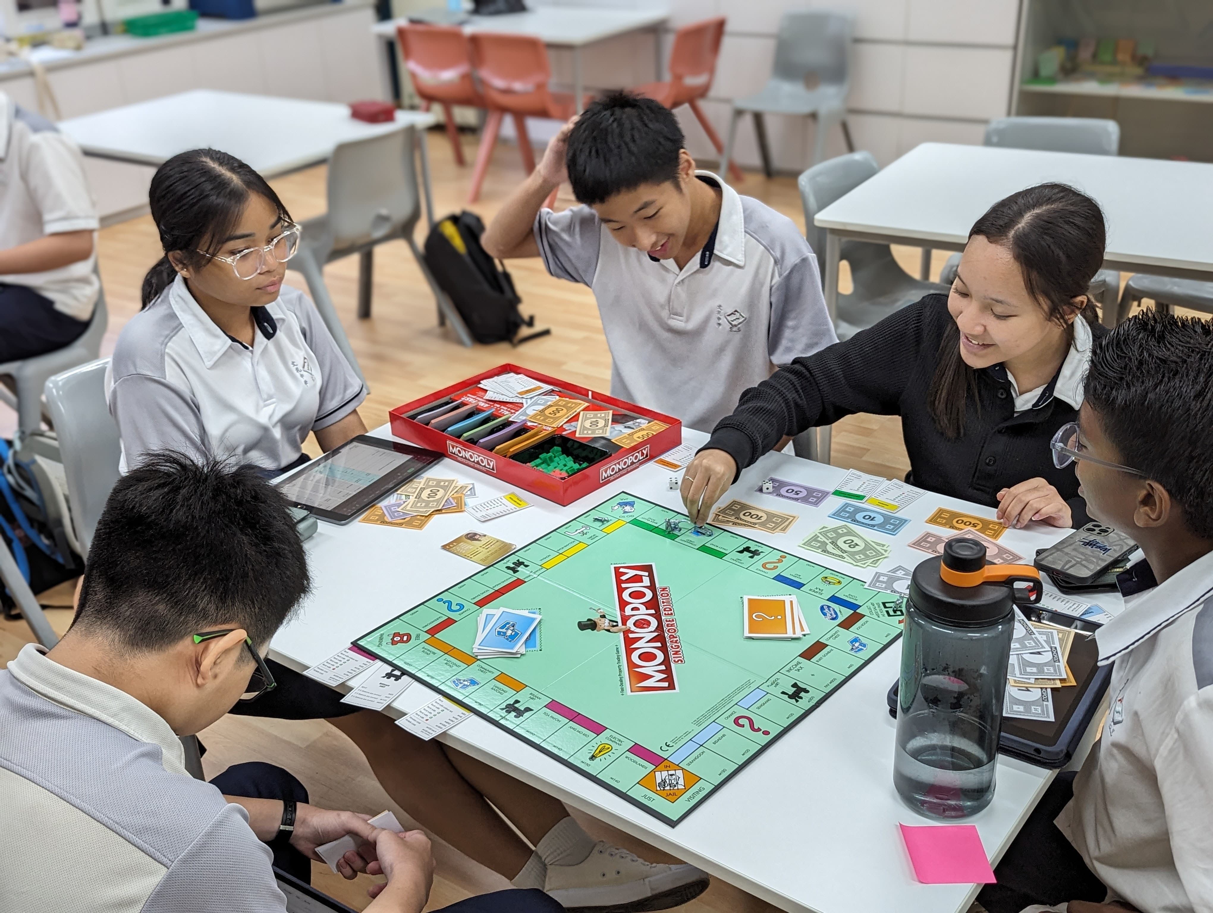 Four students in school uniforms play Monopoly Singapore Edition at a classroom table with play money and cards.