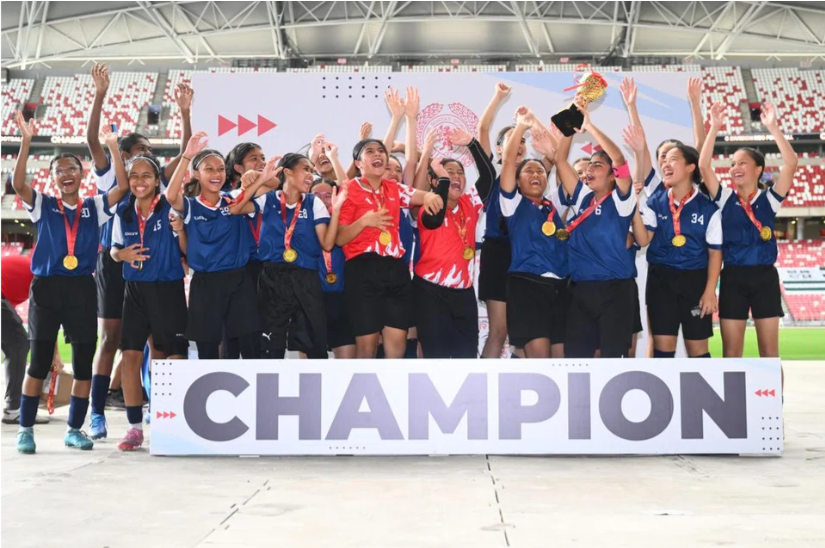 Girls football team celebrating with gold medals and a trophy on a stage with a Champion banner.