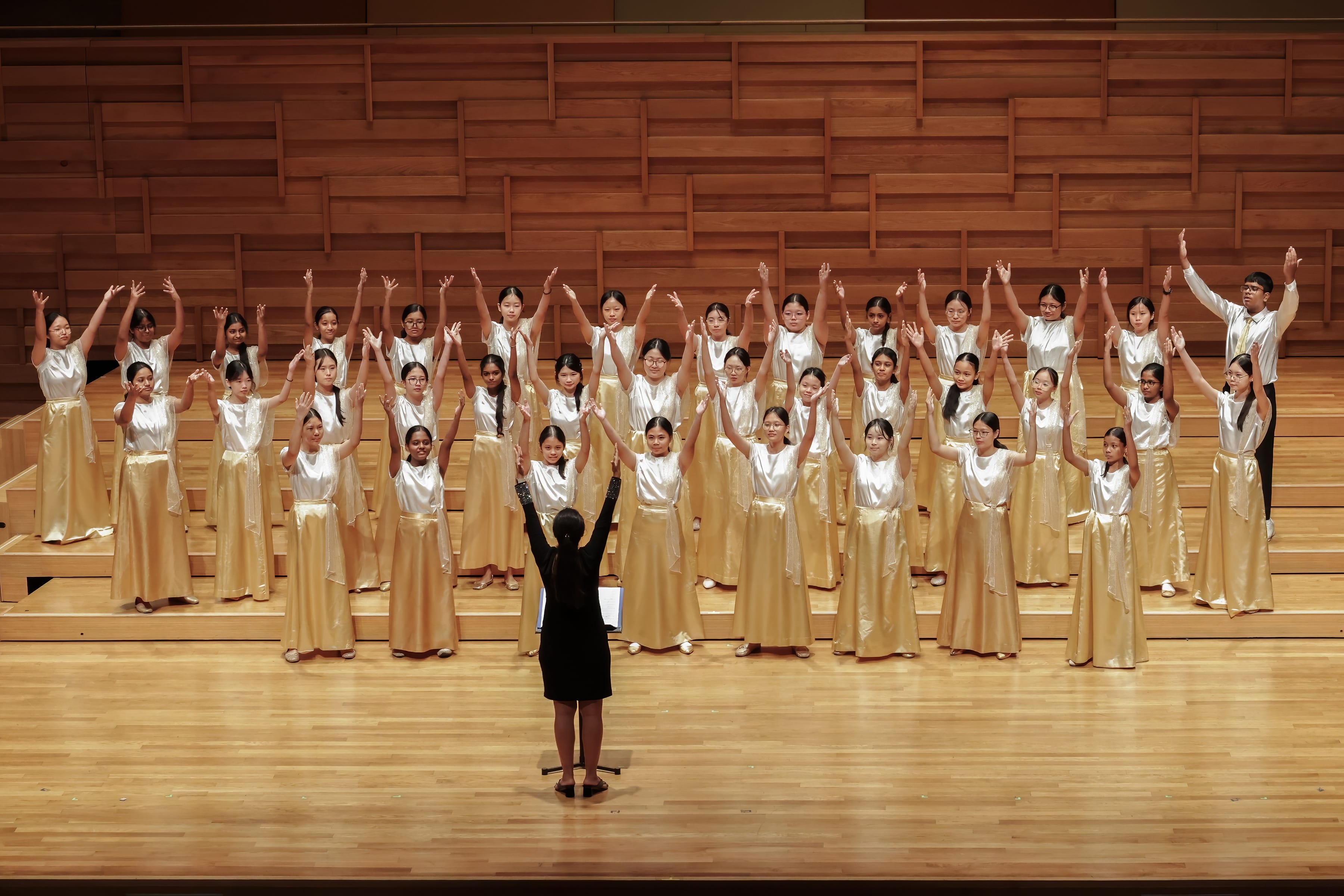 A conductor faces a large youth choir in white and gold gowns on a wooden stage, all with arms raised.