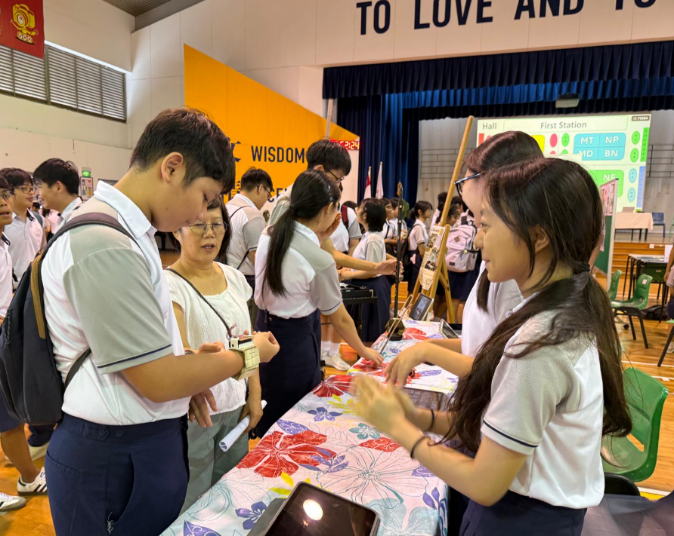 Students in white uniforms gather around a floral-covered table at a school fair inside a gymnasium.