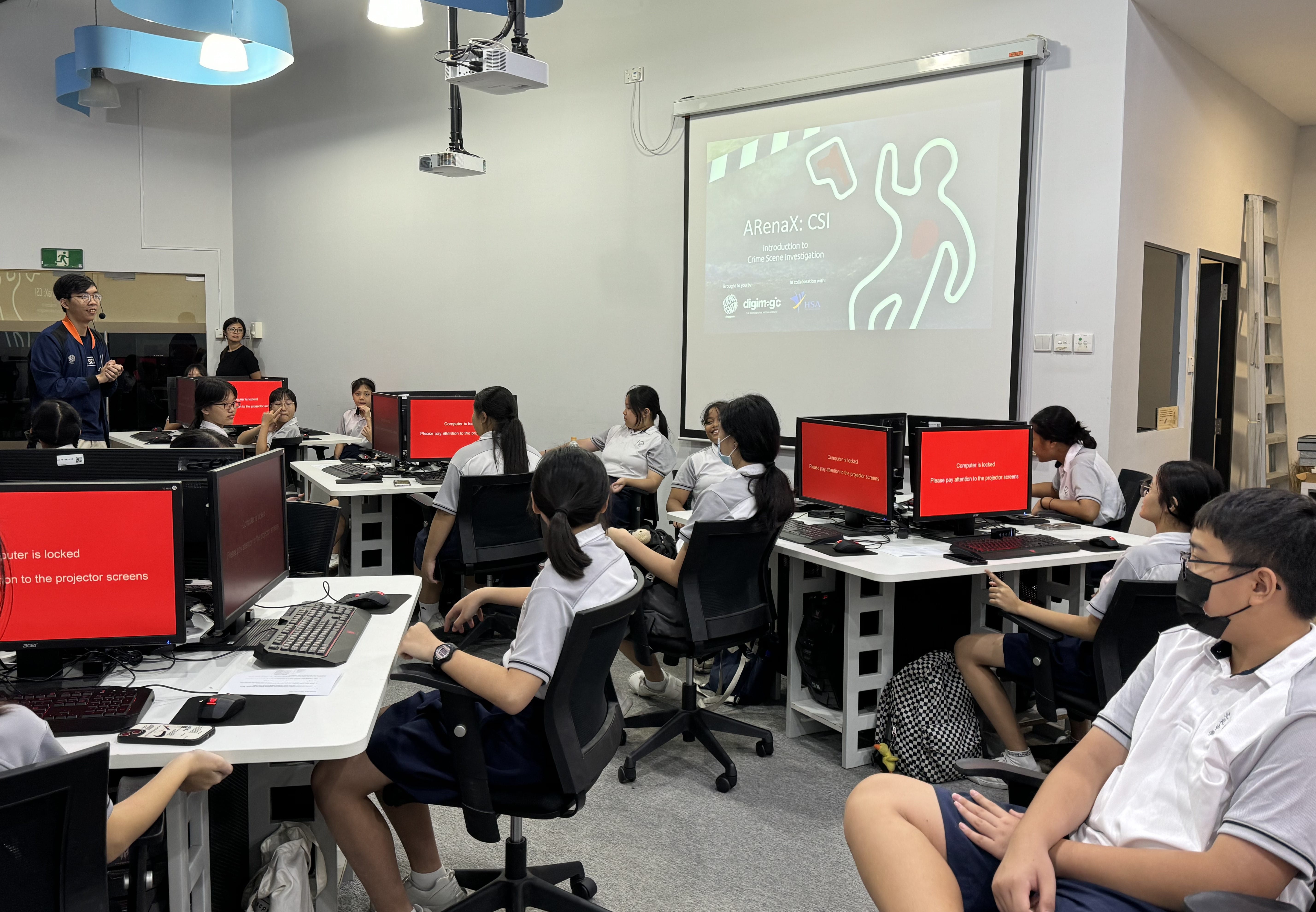 Students in a computer lab watch a projected ARenaX CSI crime scene investigation presentation, with red locked screens on desks.