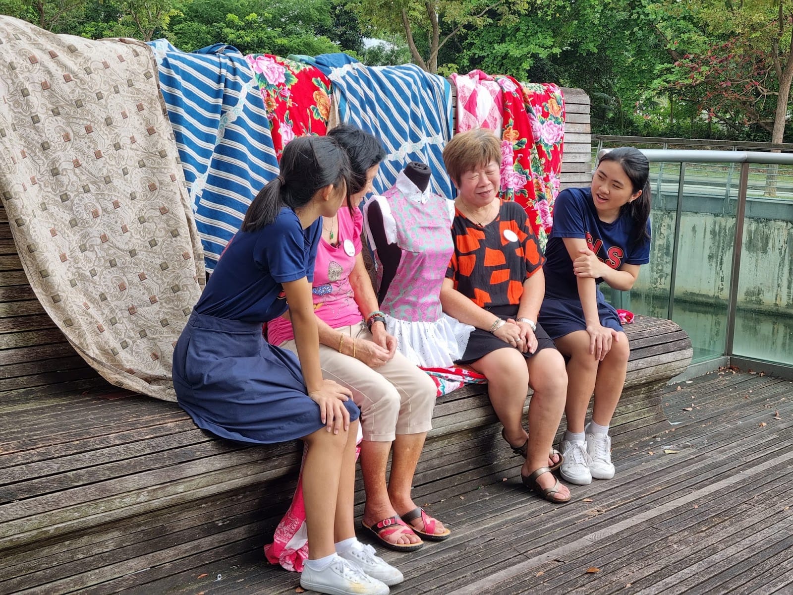 Four women sit on a wooden deck, chatting near colorful hanging fabrics and a dress mannequin, with trees behind.