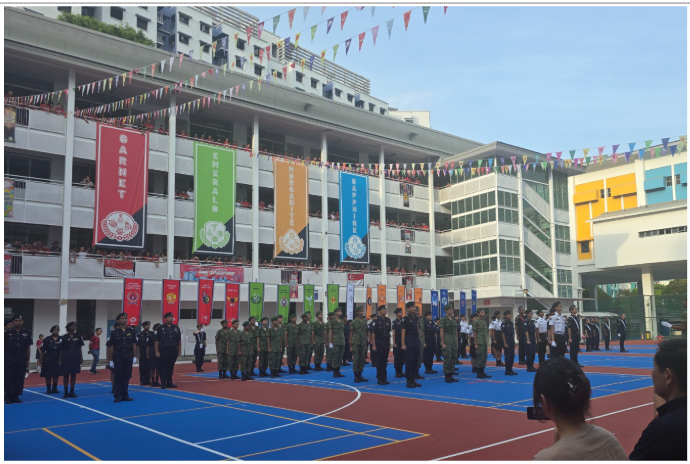Uniformed students stand in rows on a colorful court during a school ceremony with large banners and bunting.