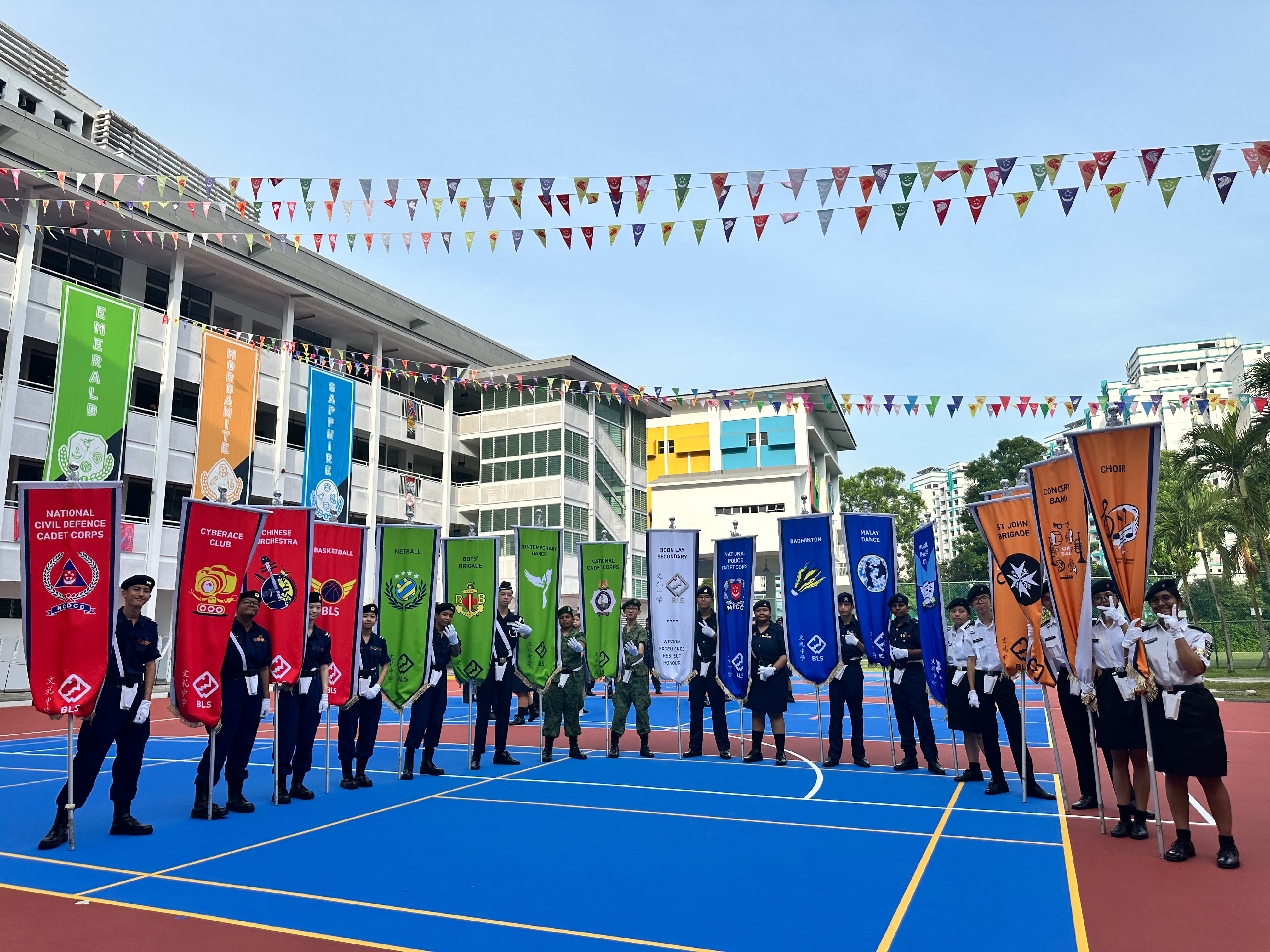 Students in uniform hold colorful club and cadet banners on a blue school court decorated with bunting.