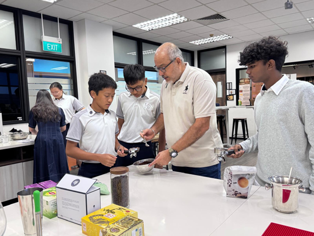 An adult man guides three school students around a white counter with coffee-making equipment in a classroom.