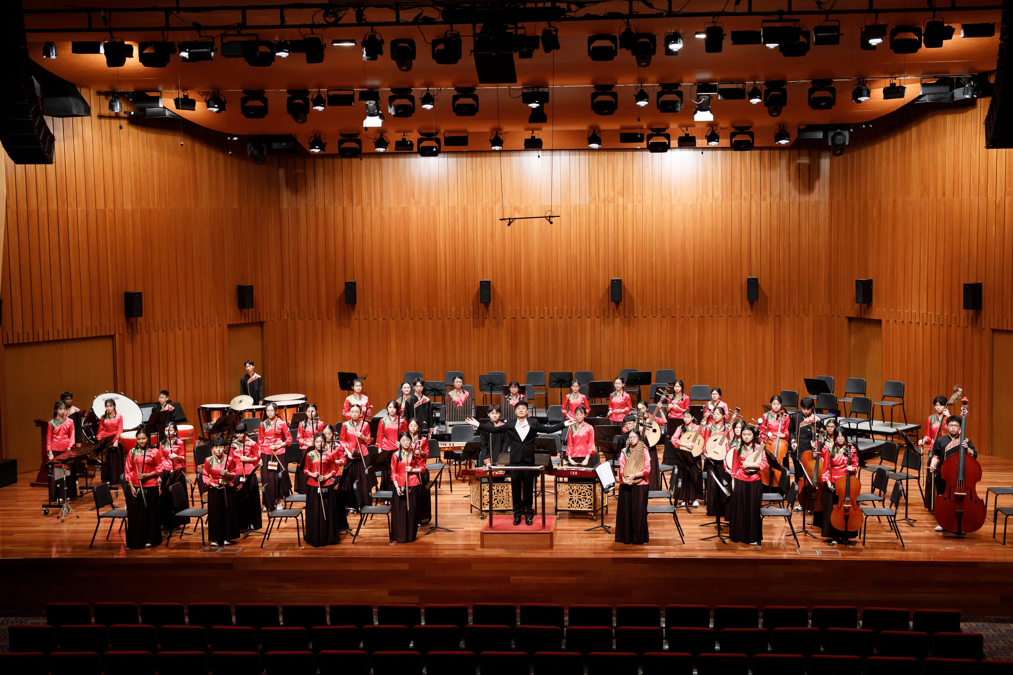 A large Chinese orchestra in red and black attire stands on a wooden concert stage with a conductor at center.