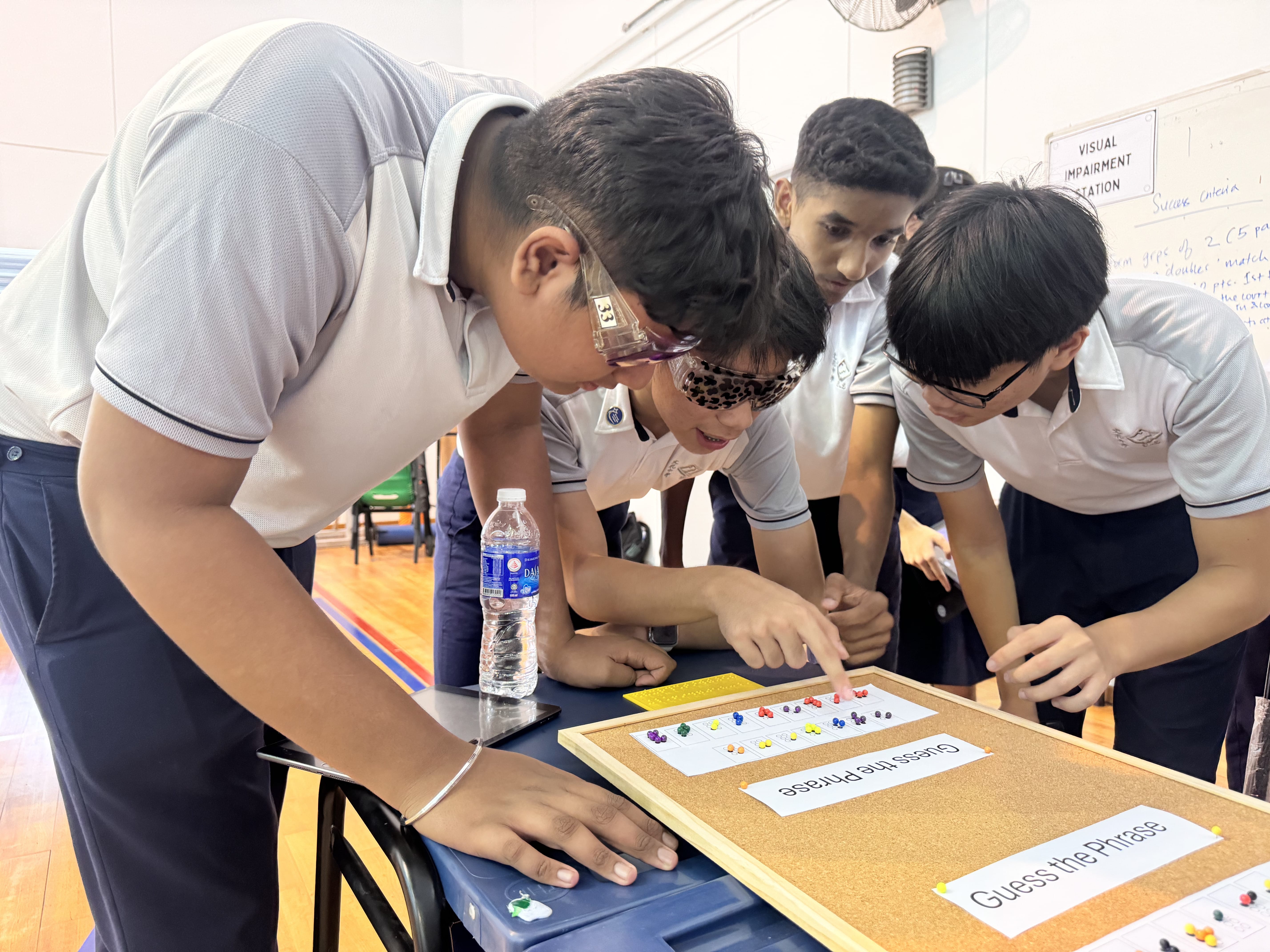 Four students in uniforms lean over a table, pointing at a pinboard game with a blindfolded student.