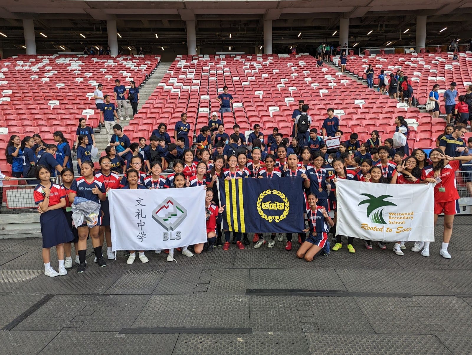 Large group of students posing with school flags from BLS, MGS, and Westwood Secondary in a stadium with red seats.