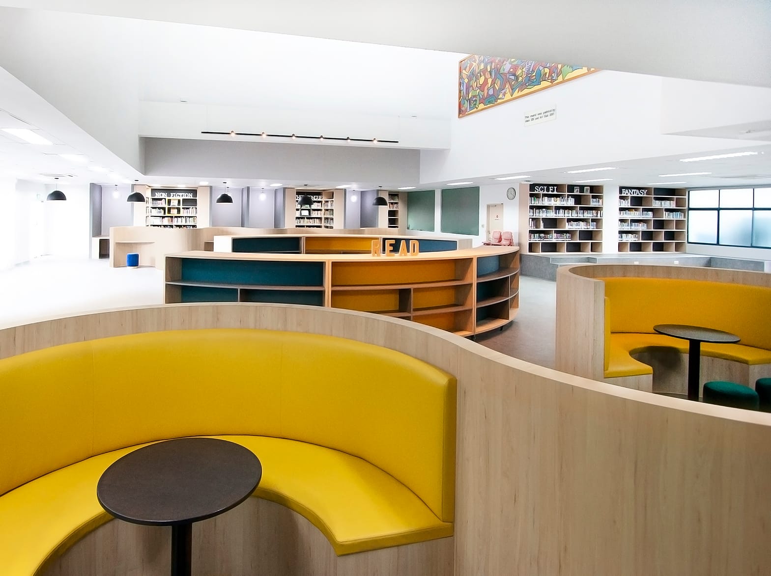 Modern library interior with curved yellow booth seating, wooden shelving units, and bookshelves labeled Sci-Fi and Fantasy.