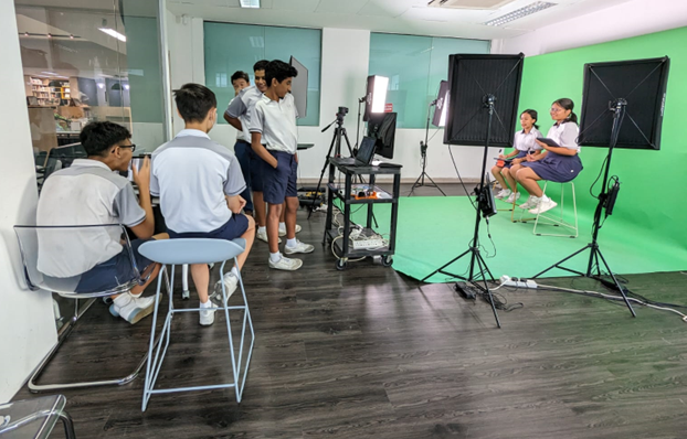 Students in school uniforms film a video production with studio lights and a green screen backdrop.